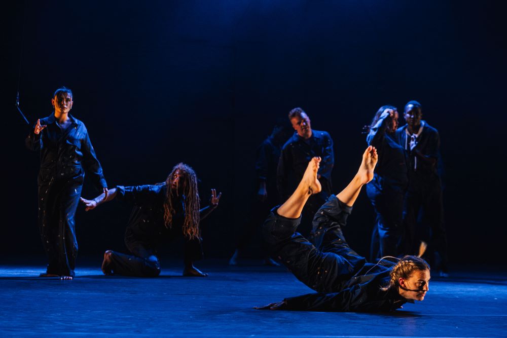 Roots compagnie 2022 danser op de voorgrond op de buik, vier andere in gelijkend uniform op de achtergrond