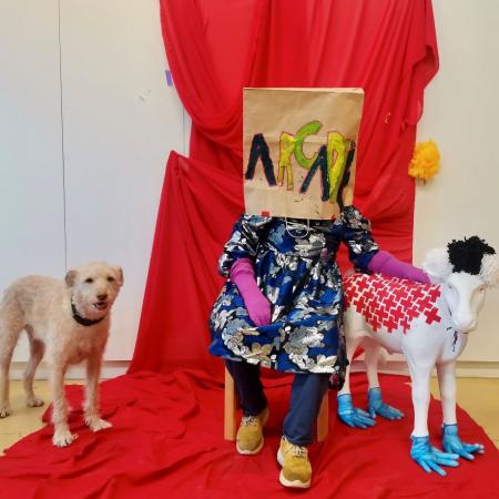 Promobeeld Arcadië kunstenares zit met papieren tas over het hoofd op een stoel voor een rood doek, links een hond, rechts een even grote replica van een schaap