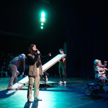 Circus zonder Handen_benefietavond presentatrice op de voorgrond, op de achtergrond bereiden twee mannen een circusact voor, rechts van haar zie je de tweede presentatrice die naar het publiek wijst