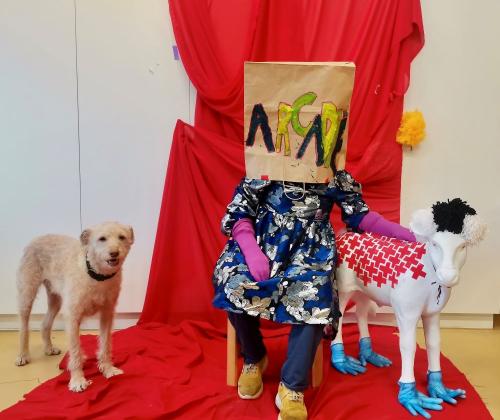 Promobeeld Arcadië kunstenares zit met papieren tas over het hoofd op een stoel voor een rood doek, links een hond, rechts een even grote replica van een schaap