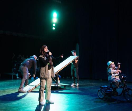 Circus zonder Handen_benefietavond presentatrice op de voorgrond, op de achtergrond bereiden twee mannen een circusact voor, rechts van haar zie je de tweede presentatrice die naar het publiek wijst